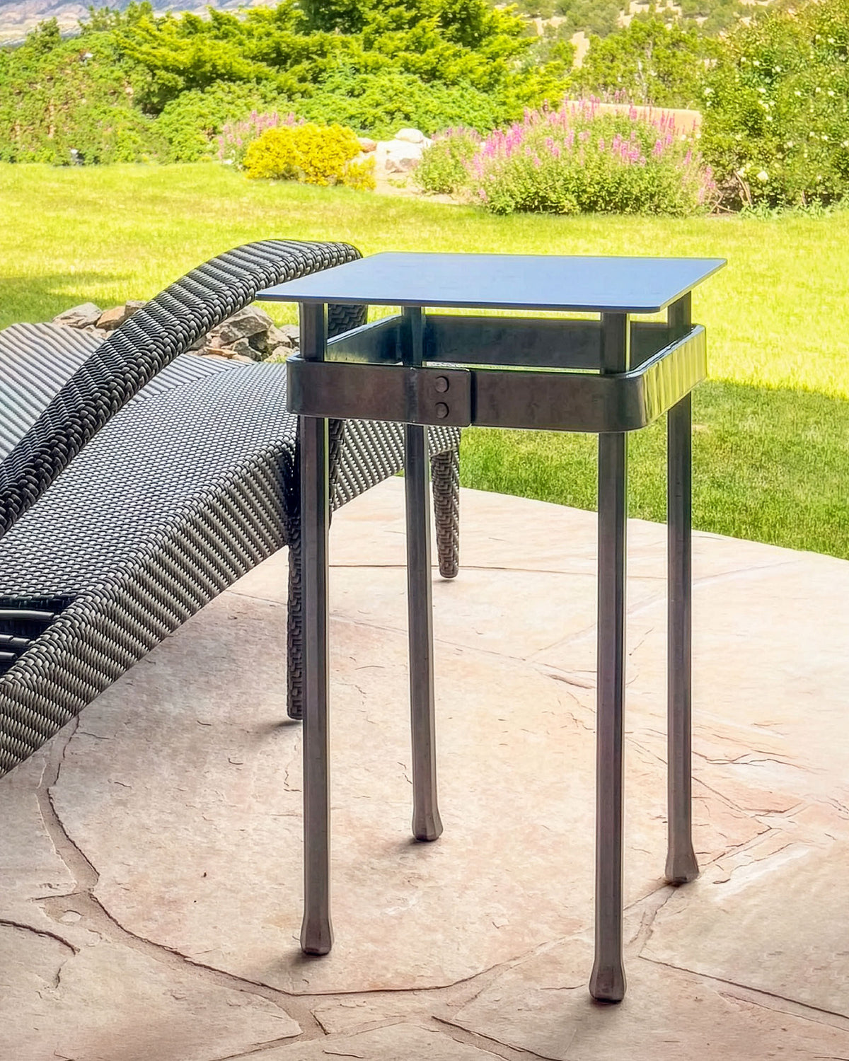 Wrought iron pedestal table with a square steel top and riveted forged band, beside a woven lounge chair on a stone patio