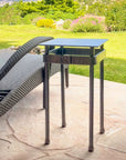 Wrought iron pedestal table with a square steel top and riveted forged band, beside a woven lounge chair on a stone patio