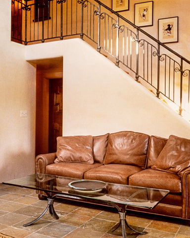 A sitting area with a wrought iron coffee table and a brown leather couch positioned just in front of a staircase with a custom wrought iron staircase railing. 