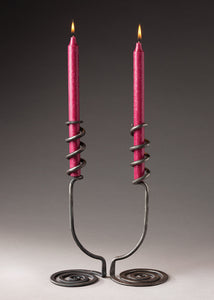 Two artistic hand forged candlesticks with a swirling base that reaches upwards and wraps around the base of the red candles. 