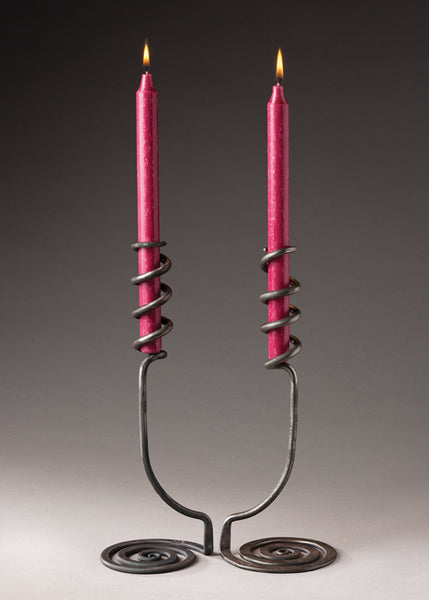 Two artistic hand forged candlesticks with a swirling base that reaches upwards and wraps around the candles. 
