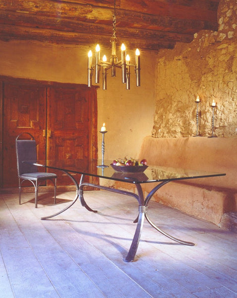 A rustic, elegantly decorated dining area w/ a long, forged steel dinner party table with a glass top in the center of the room. Hanging from the wooden beam ceiling is a large wrought iron chandelier with 6 tall torches with candelabra light bulbs. Tall forged steel candlestick holders decorate the table and the adobe walls.