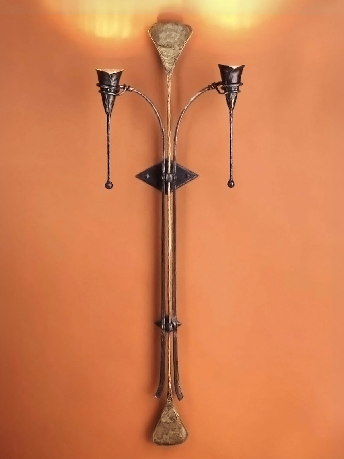  A large hand forged statement light fixture, Iron Calla Torchiere, with a long skinny central piece that flares at the top and bottom with two calla lily torches on either side mounted to an an warm toned wall