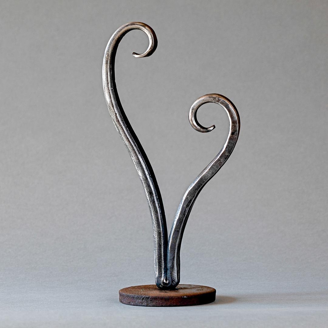 Opening Heart Sculpture made from hand forged steel. This small sculpture has a round steel base holding a heart shape sculpture that remains open at the top with each side of the heart curling inwards.