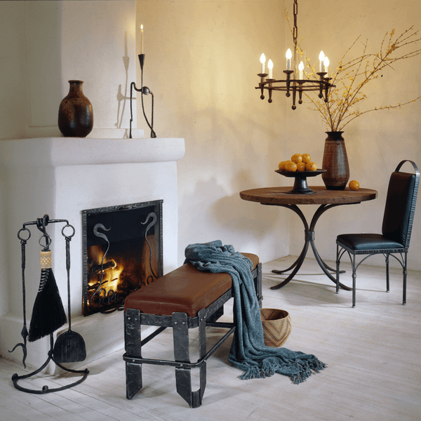 Fireside living room scene furnished by Christopher Thomson Ironworks featuring a fireplace tool set, bench, hanging chandelier, table and chair. 
