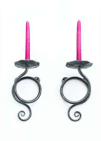 Hand forged wrought iron wall candlestick holders that hold a standard taper candle. Beneath the candle, a vine like design gracefully loops around itself .